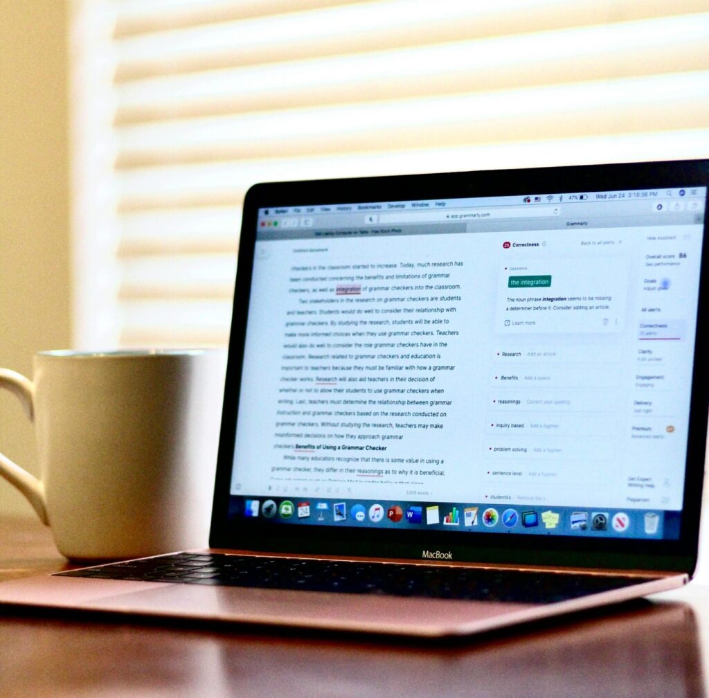 A laptop on a wooden table next to a ceramic mug, illuminated by window blinds.