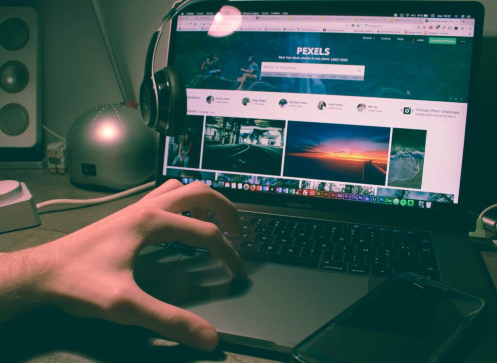 Person browsing Pexels on a laptop with headphones and smartphone nearby.